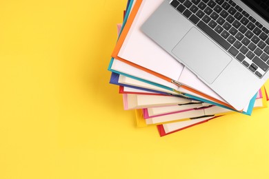 Stack of colorful books and laptop on yellow background, top view. Space for text