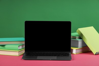 Many colorful books and laptop on pink table against green background