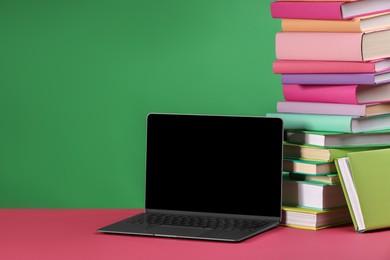 Photo of Stack of colorful books and laptop on pink table against green background, space for text