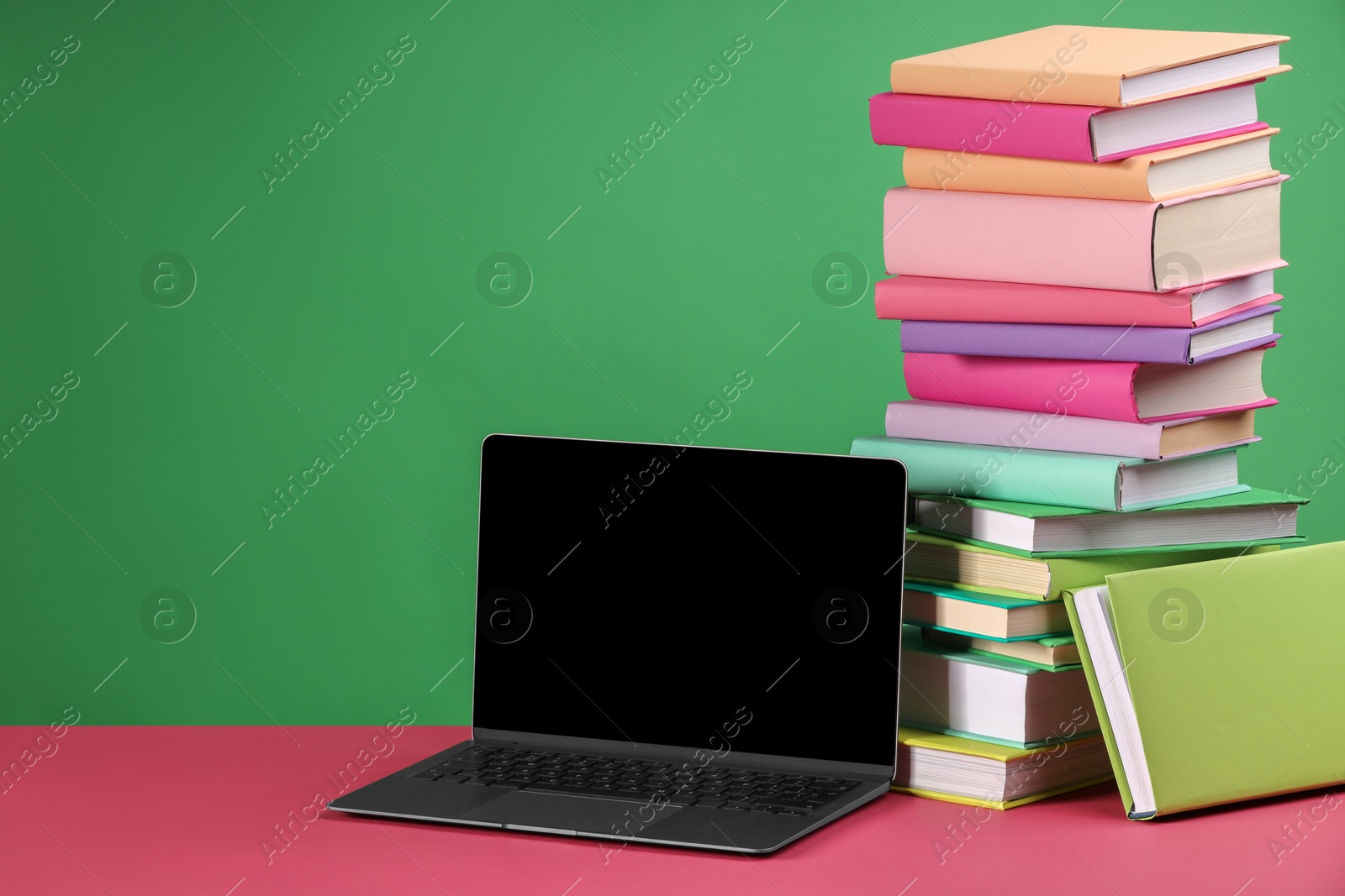 Photo of Stack of colorful books and laptop on pink table against green background, space for text