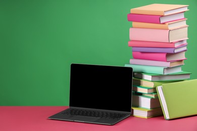 Photo of Stack of colorful books and laptop on pink table against green background, space for text