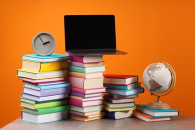 Photo of Stacks of colorful books, globe, clock and laptop on wooden table against orange background