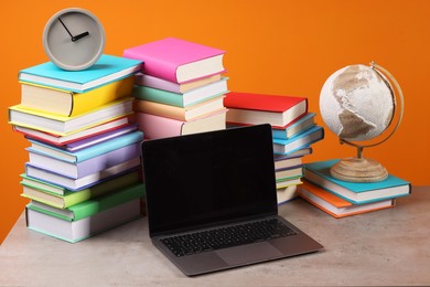 Photo of Stacks of colorful books, globe, clock and laptop on wooden table against orange background