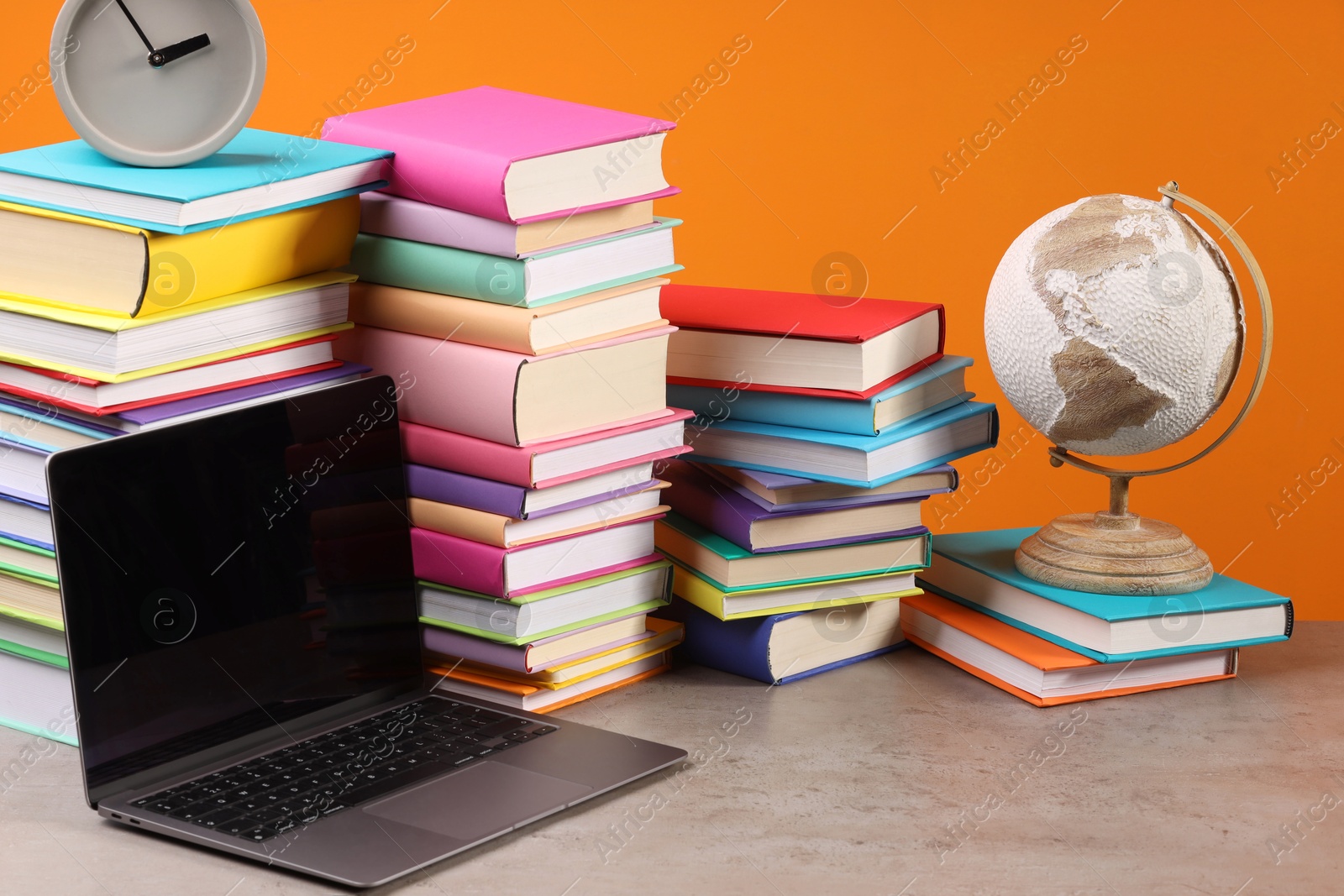 Photo of Stacks of colorful books, globe, clock and laptop on wooden table against orange background