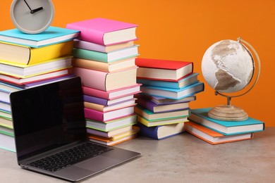 Photo of Stacks of colorful books, globe, clock and laptop on wooden table against orange background