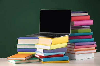 Stacks of many colorful books and laptop on white table near chalkboard in classroom