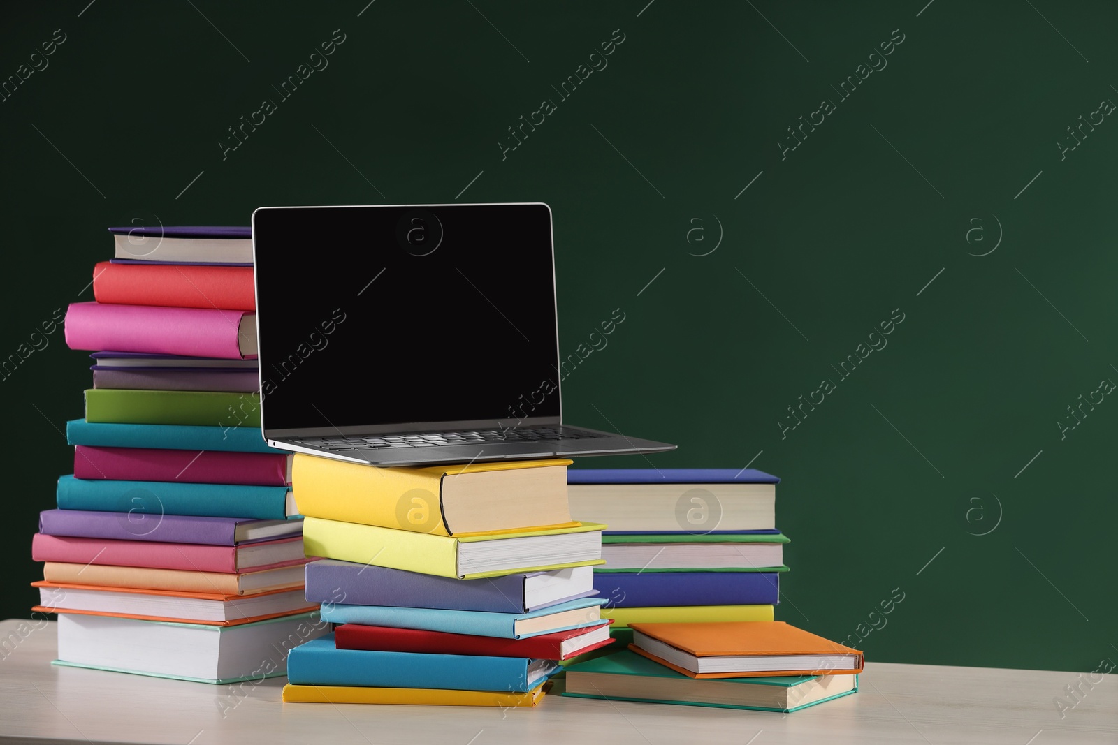 Photo of Stacks of many colorful books and laptop on white table near chalkboard in classroom. Space for text