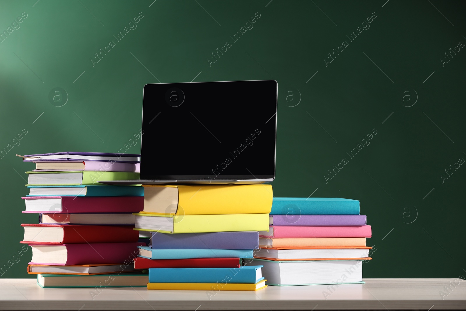 Photo of Stacks of many colorful books and laptop on white wooden table near chalkboard in classroom