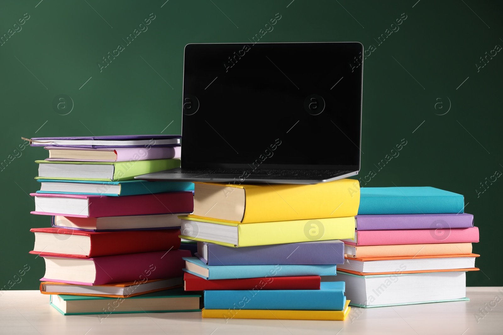 Photo of Stacks of many colorful books and laptop on white wooden table near chalkboard in classroom