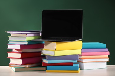 Photo of Stacks of many colorful books and laptop on white wooden table near chalkboard in classroom