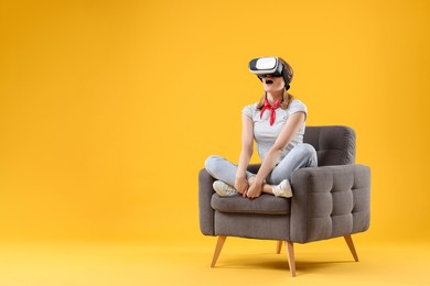 Emotional woman with virtual reality headset sitting in armchair on yellow background, space for text