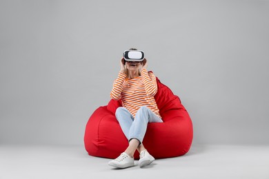 Happy woman with virtual reality headset sitting on bean bag chair against gray background