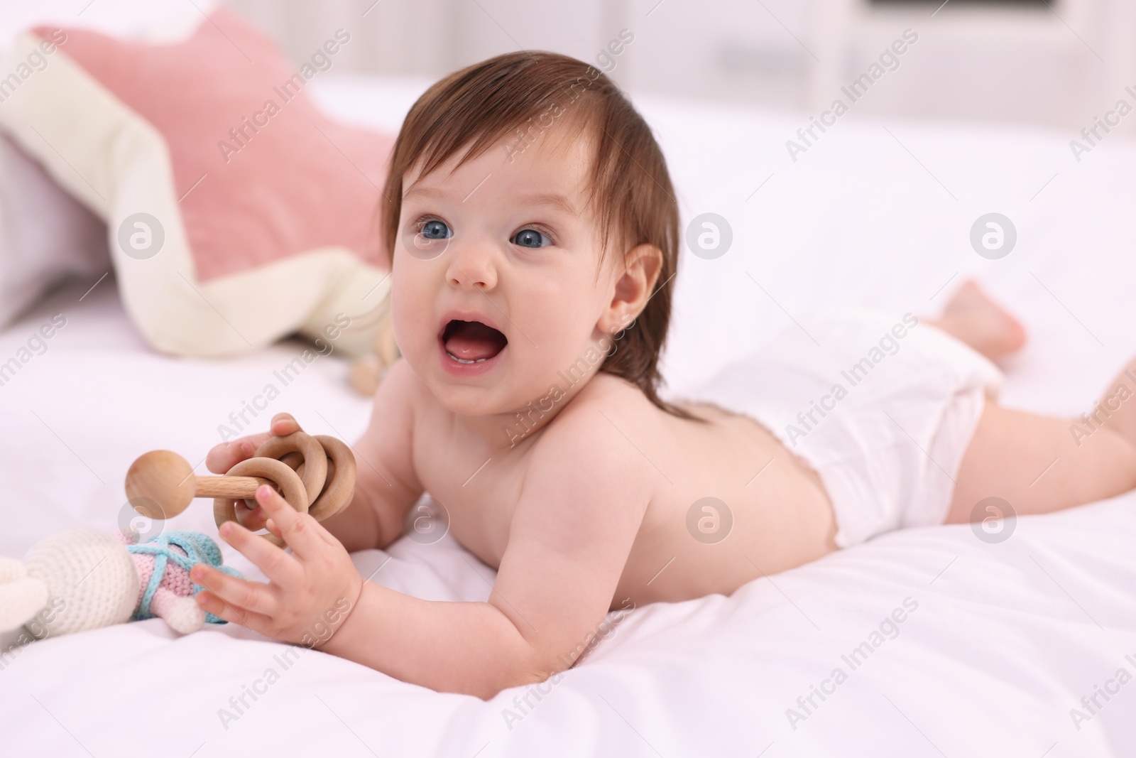 Photo of Cute little girl in diaper with wooden teether and toy bunny on bed at home