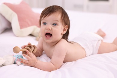 Photo of Cute little girl in diaper with wooden teether and toy bunny on bed at home