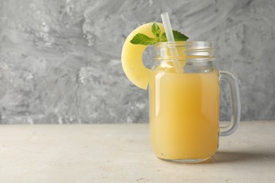 Photo of Tasty pineapple juice in mason jar, mint and slice of fresh fruit on grey textured table, closeup. Space for text