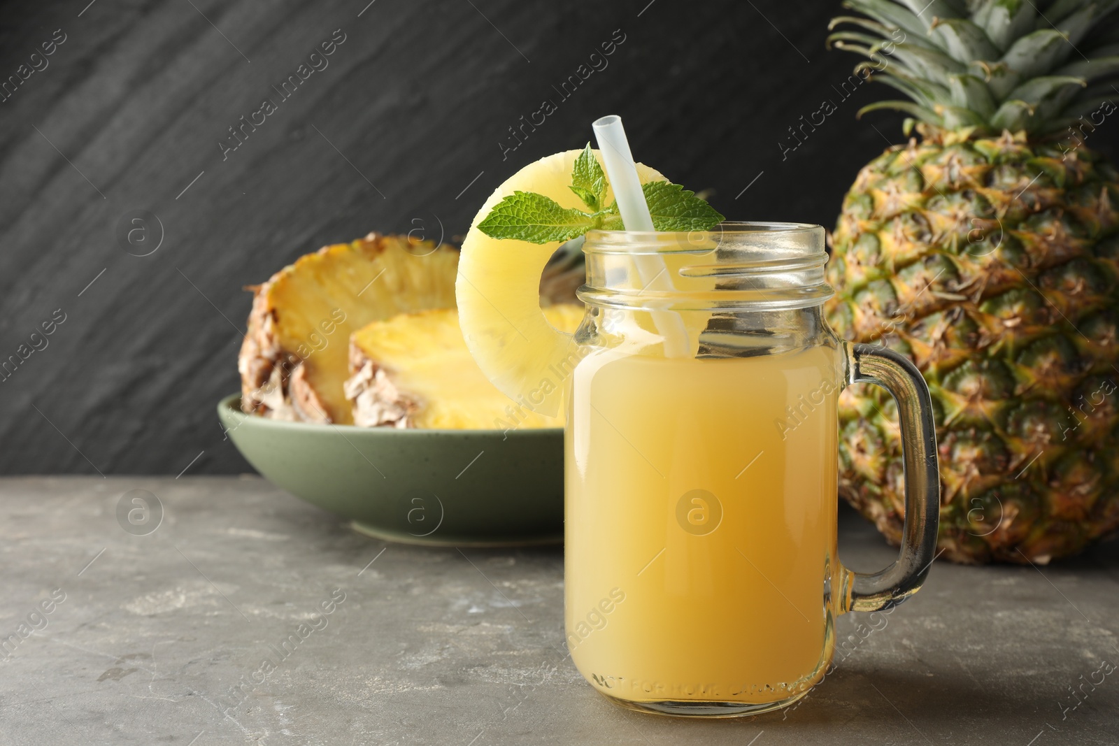 Photo of Tasty pineapple juice in mason jar, mint and slice of fruit on grey textured table, closeup. Space for text