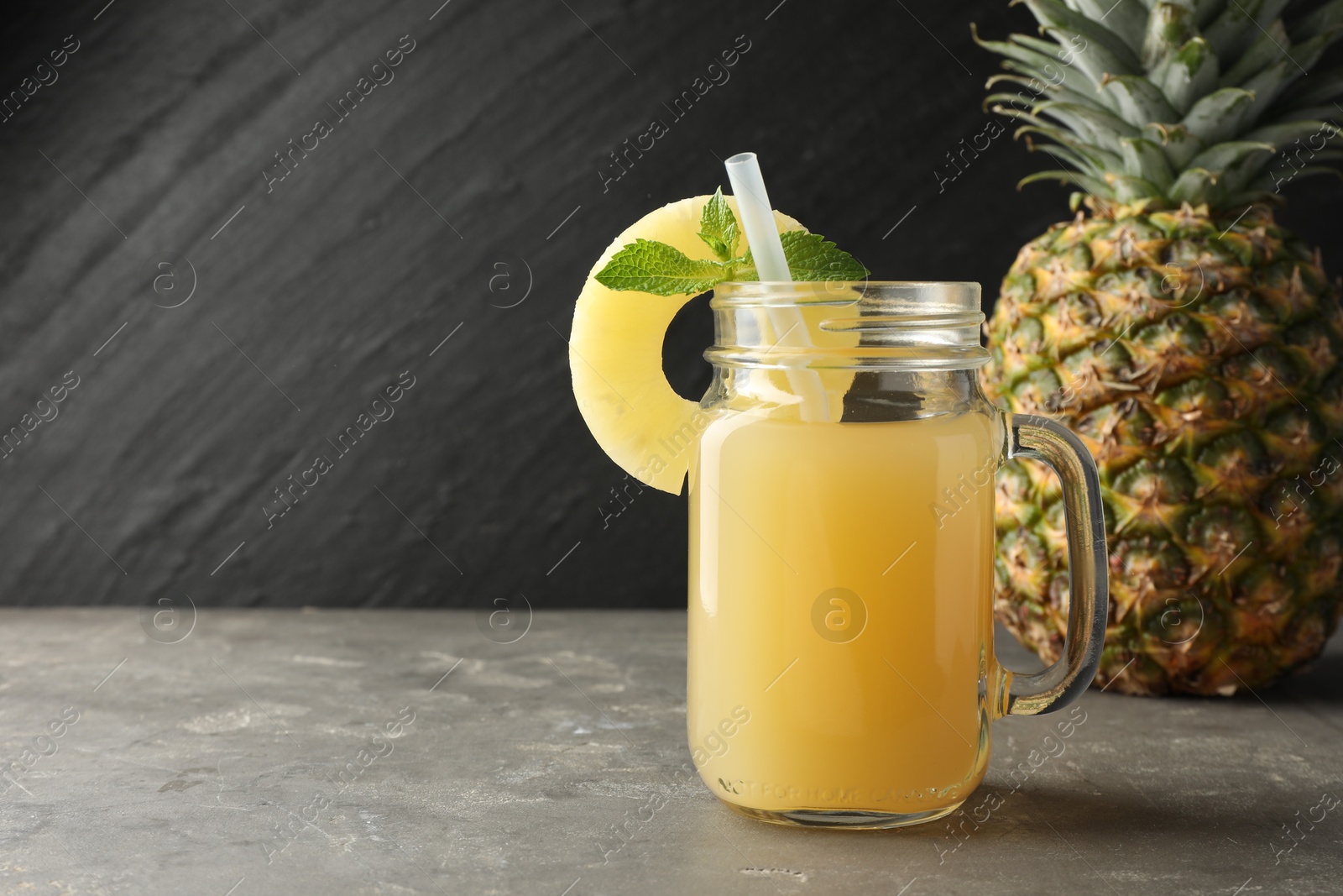 Photo of Tasty pineapple juice in mason jar, mint and slice of fruit on grey textured table, closeup. Space for text