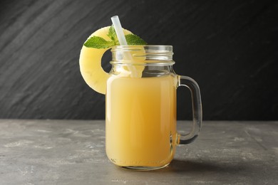 Photo of Tasty pineapple juice in mason jar, mint and slice of fruit on grey textured table, closeup