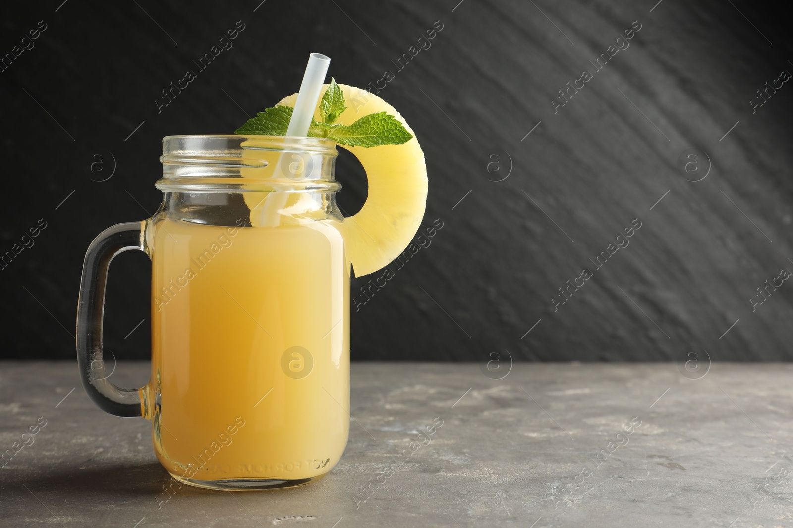 Photo of Tasty pineapple juice in mason jar, mint and slice of fruit on grey textured table, closeup. Space for text