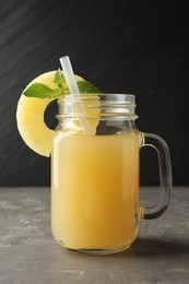 Photo of Tasty pineapple juice in mason jar, mint and slice of fruit on grey textured table, closeup