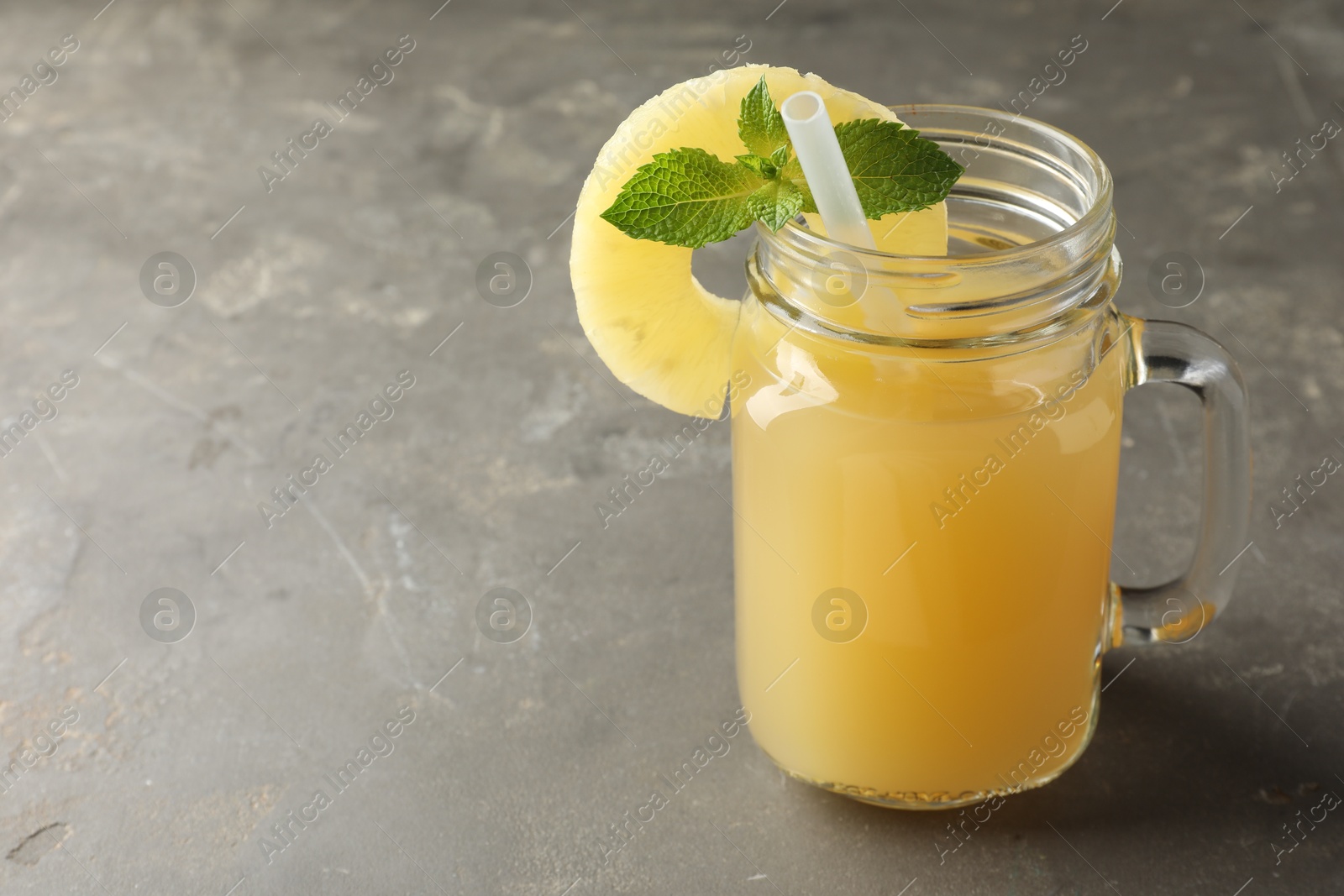 Photo of Tasty pineapple juice in mason jar, mint and slice of fruit on grey textured table, closeup. Space for text