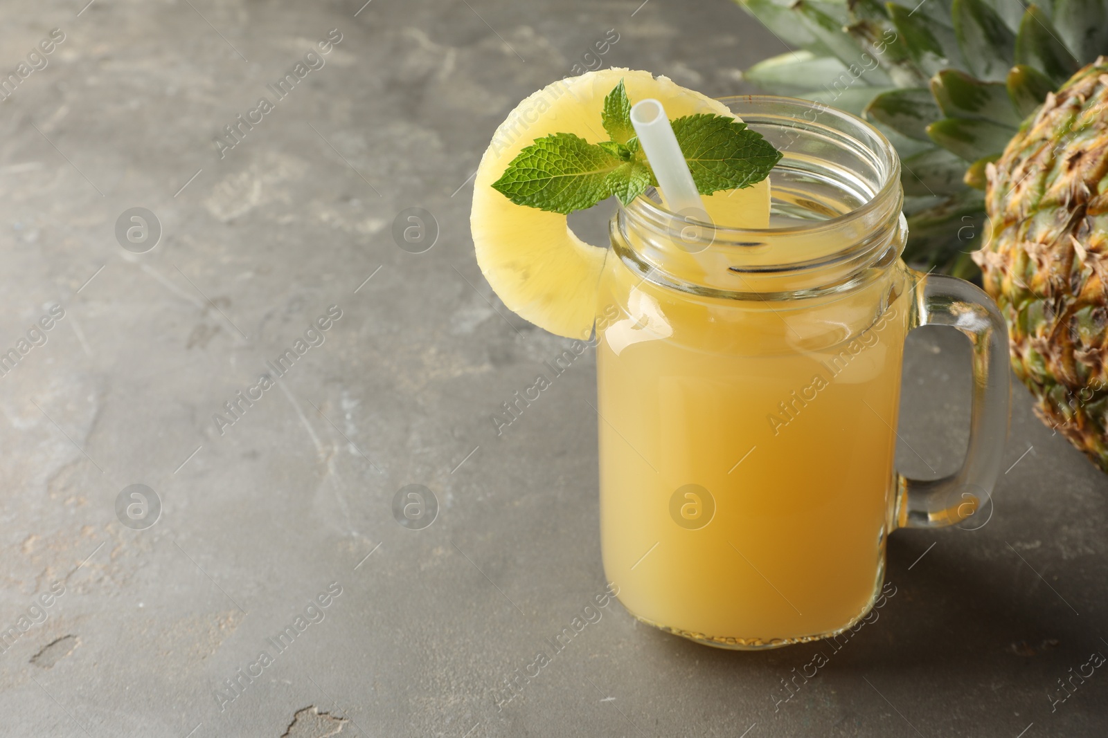 Photo of Tasty pineapple juice in mason jar, mint and slice of fruit on grey textured table, closeup. Space for text