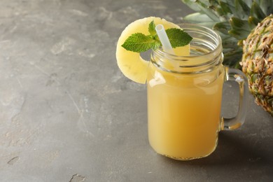 Photo of Tasty pineapple juice in mason jar, mint and slice of fruit on grey textured table, closeup. Space for text