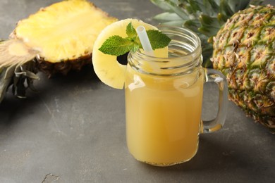 Photo of Tasty pineapple juice in mason jar, mint and fruits on grey textured table, closeup