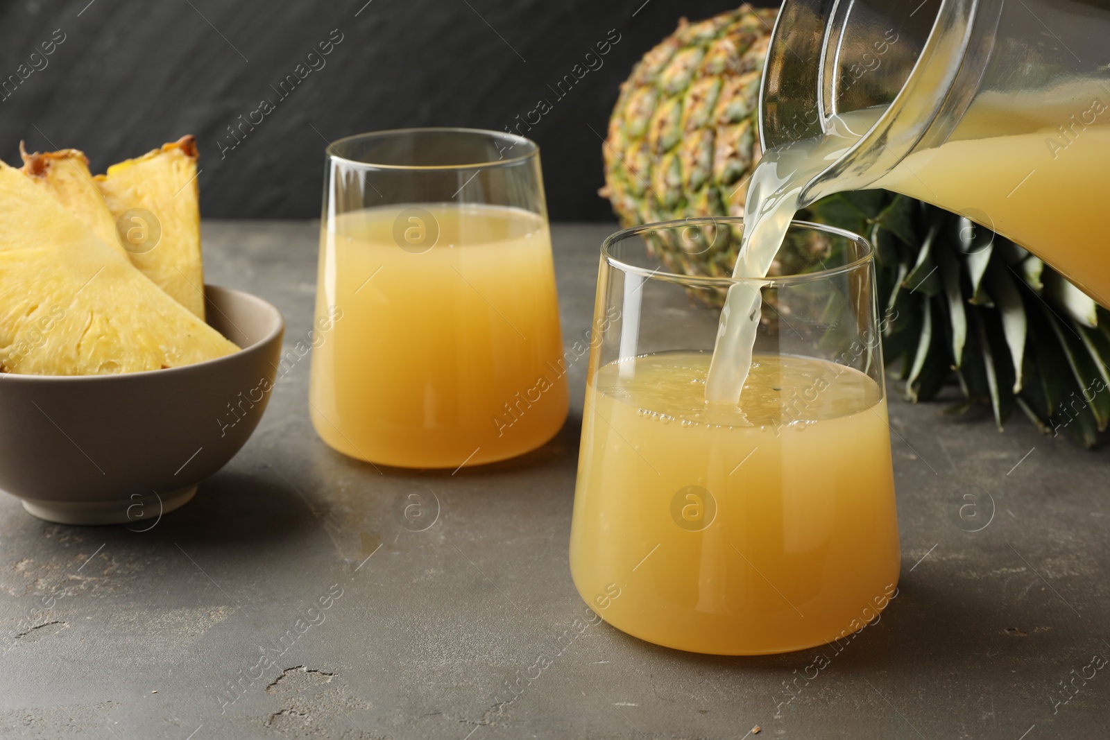 Photo of Pouring tasty pineapple juice from jar into glass at grey textured table, closeup