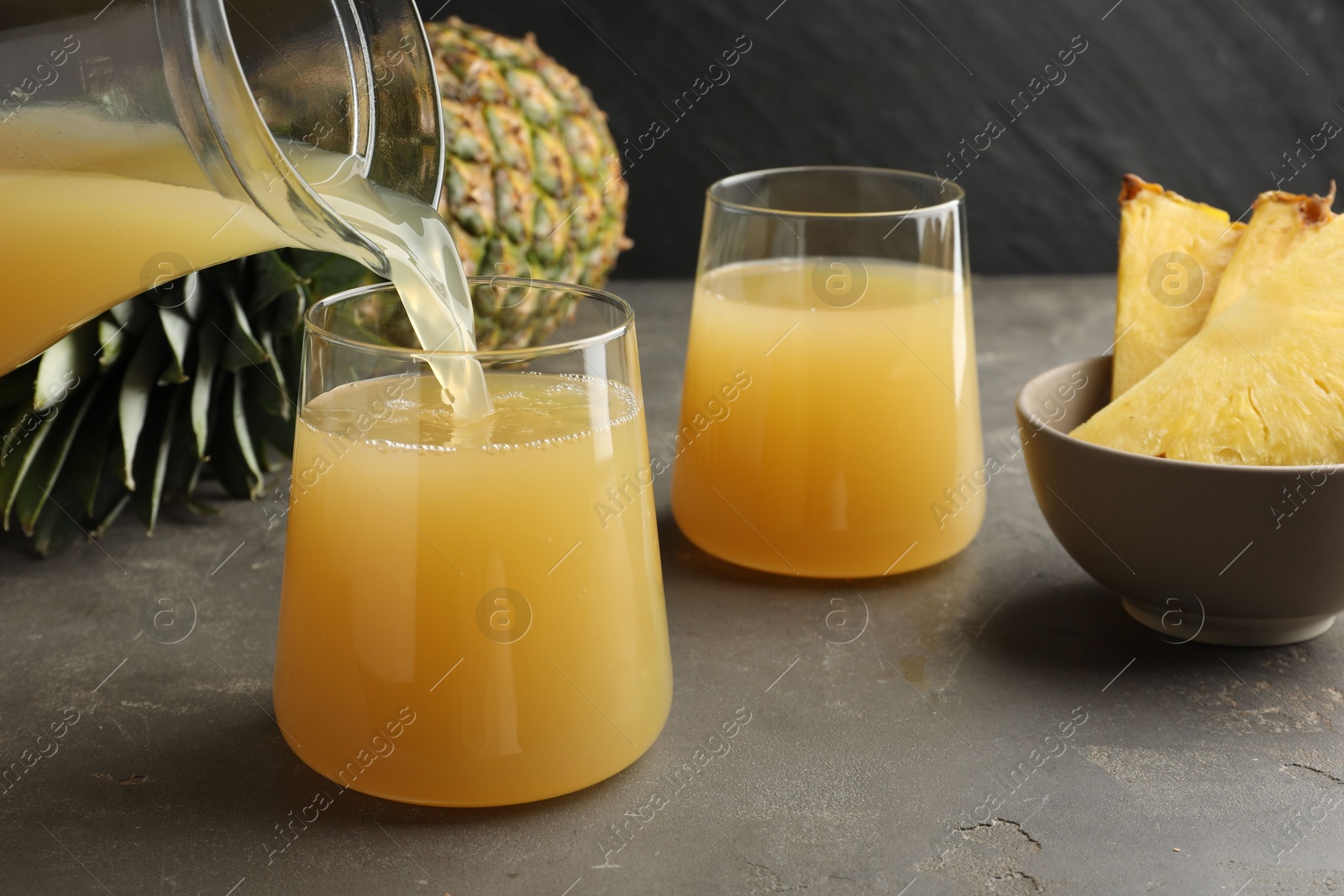 Photo of Pouring tasty pineapple juice from jar into glass at grey textured table, closeup