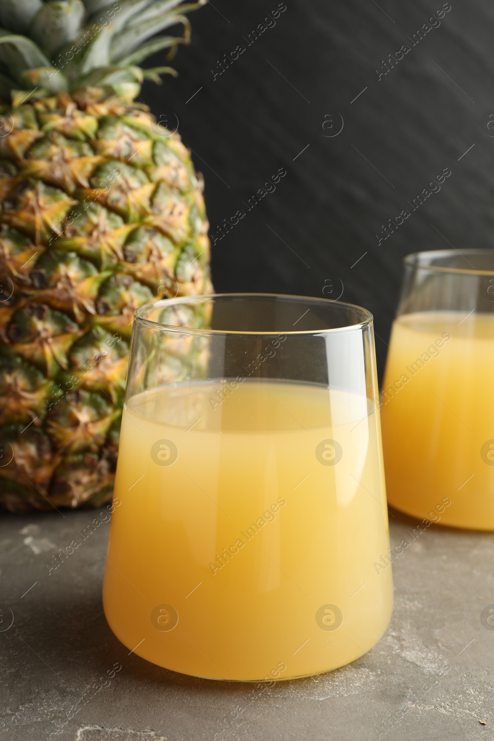 Photo of Tasty pineapple juice in glass and fruits on grey textured table, closeup