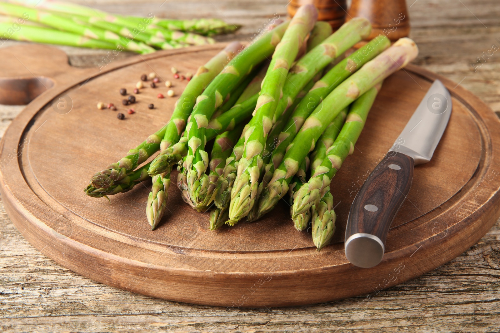 Photo of Board with fresh green asparagus stems, peppercorns and knife on wooden table, closeup