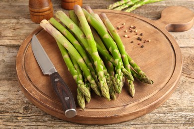 Photo of Board with fresh green asparagus stems, peppercorns and knife on wooden table, closeup