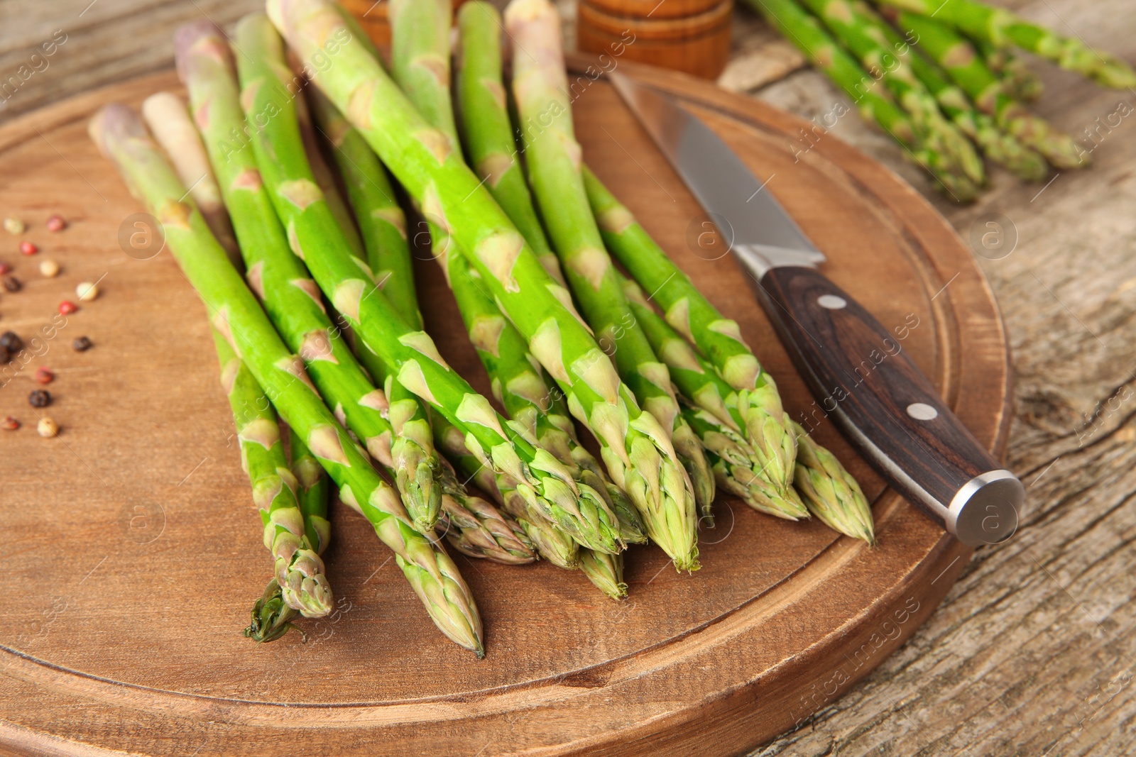 Photo of Board with fresh green asparagus stems, peppercorns and knife on wooden table, closeup