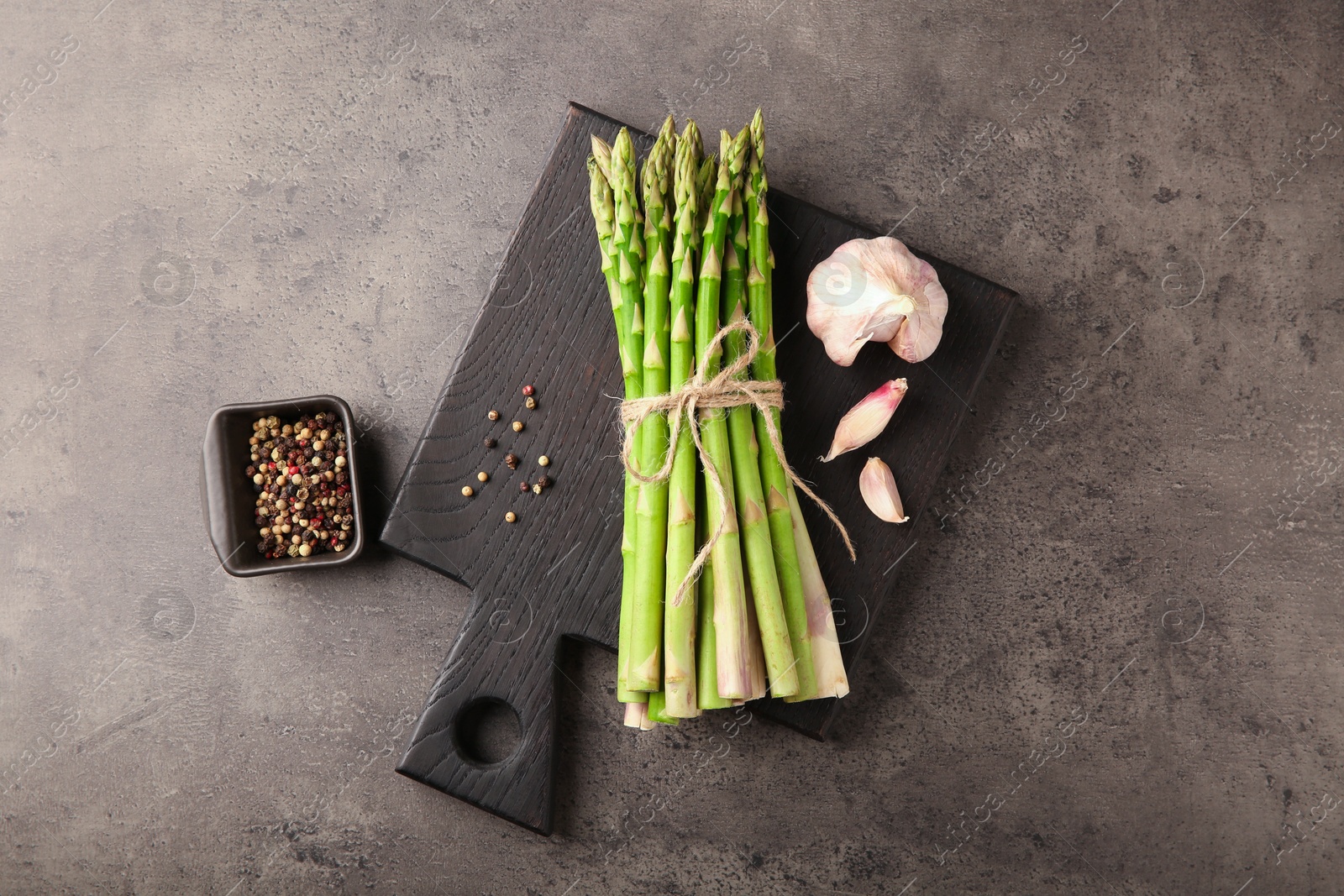 Photo of Board with bunch of fresh green asparagus stems, garlic and peppercorns on grey textured table, flat lay