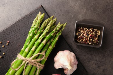 Photo of Board with bunch of fresh green asparagus stems, garlic and peppercorns on grey textured table, flat lay