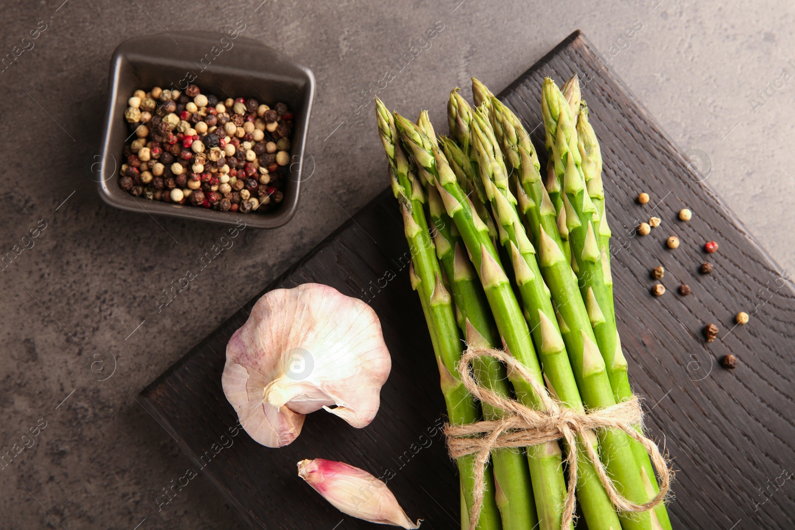 Photo of Board with bunch of fresh green asparagus stems, garlic and peppercorns on grey textured table, flat lay