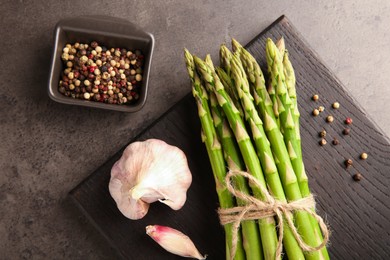 Board with bunch of fresh green asparagus stems, garlic and peppercorns on grey textured table, flat lay