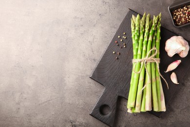 Photo of Board with bunch of fresh green asparagus stems, garlic and peppercorns on grey textured table, flat lay. Space for text