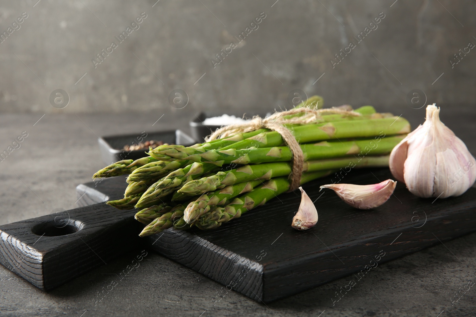 Photo of Board with bunch of fresh green asparagus stems and garlic on grey textured table, closeup