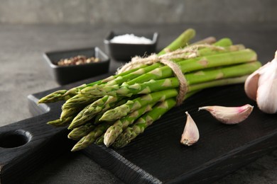 Board with bunch of fresh green asparagus stems and garlic on grey textured table, closeup