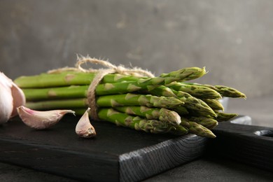 Board with bunch of fresh green asparagus stems and garlic on grey textured table, closeup