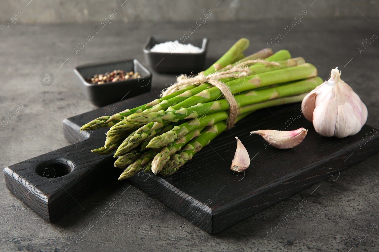 Photo of Board with bunch of fresh green asparagus stems and garlic on grey textured table, closeup