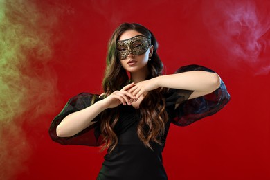 Photo of Beautiful woman wearing carnival mask on red background in color lights and smoke