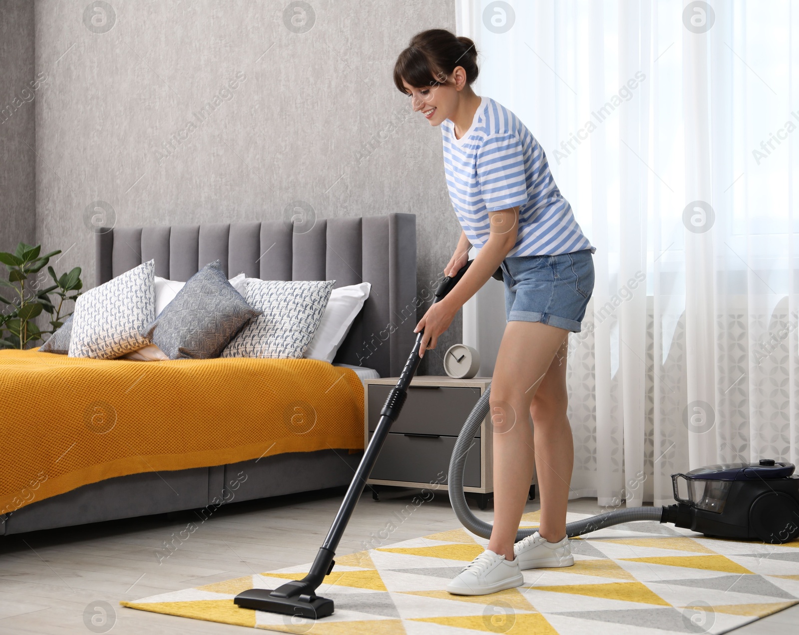 Photo of Young woman cleaning carpet with vacuum in bedroom