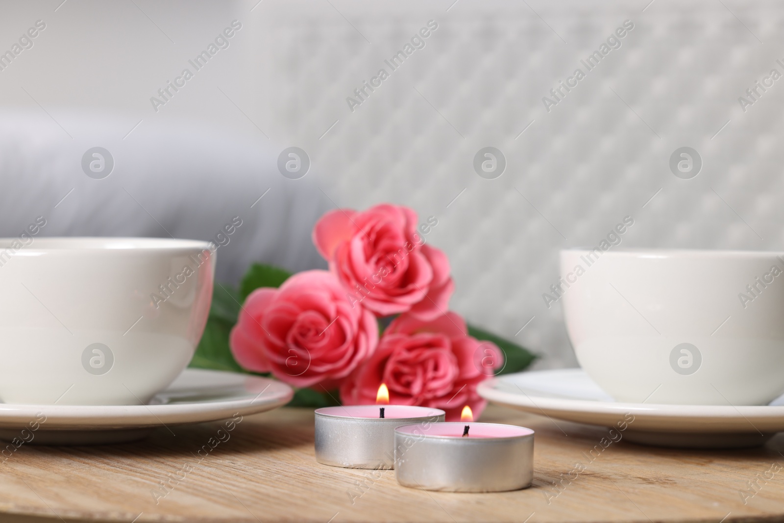 Photo of Burning candles, two cups of drink and flowers on wooden table indoors, closeup