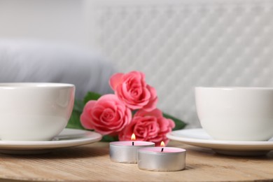 Photo of Burning candles, two cups of drink and flowers on wooden table indoors, closeup