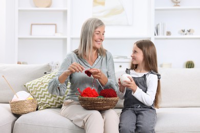 Smiling grandmother teaching her granddaughter to knit on sofa at home