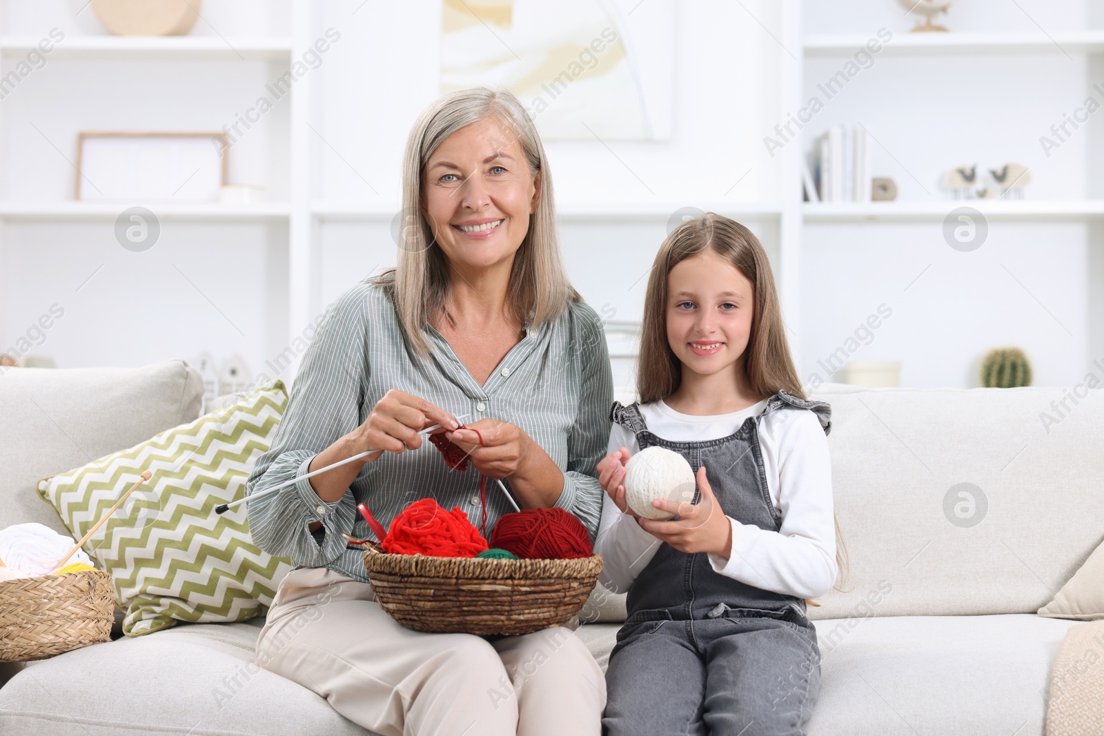 Photo of Happy grandmother with granddaughter holding knitting needles and yarn on sofa at home