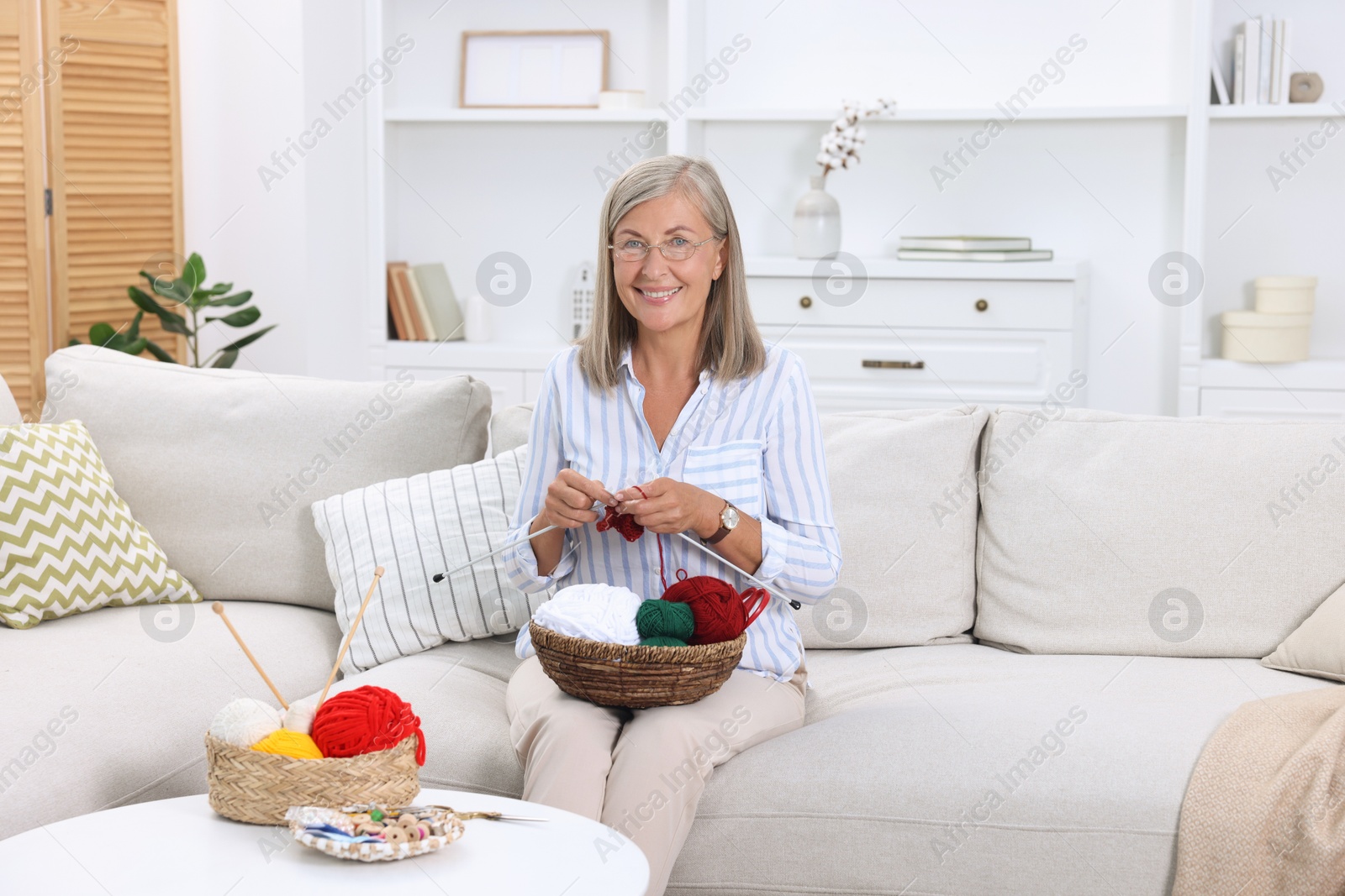 Photo of Smiling woman with basket of yarn knitting on sofa at home